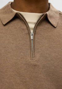 Pull en tricot marron avec un col zippé et un tissu texturé. Une couche intérieure crème est visible sous le col.