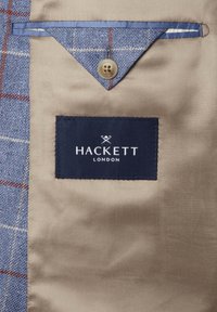 Béžová podšívka bundy s modrou károvanou klopou vrecka, upevnenou hnedým gombíkom, so štítkom značky Hackett London v čiernej farbe.