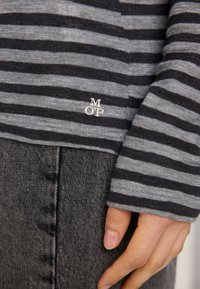 Riba-lineeritud sviiter hallide ja mustade horisontaalsete joontega, millel on peen "M OP" logo. Pehme kangas, koosneb kihilisest riietusest tume denimiga.