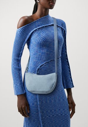 Bolso de hombro de denim azul claro con forma redondeada, cierre de cremallera y correa ajustable, que presenta un sutil logo grabado en la parte delantera.