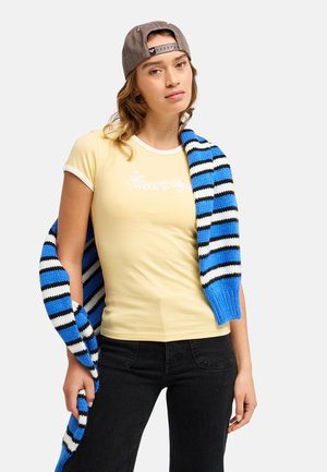 Jeune femme portant un t-shirt jaune et une casquette grise à l'envers tient une écharpe tricotée rayée bleue, noire et blanche sur l'épaule.