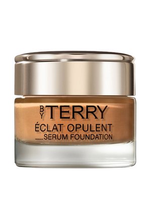 Serum foundation in een transparante glazen pot met een gouden deksel. Het product is lichtbruin met reliëf zwart tekst. Glad oppervlak, compact ontwerp.