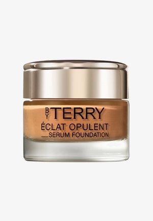 Fond de teint en sérum dans un pot en verre transparent avec un couvercle doré. Le produit est marron clair avec un texte noir en relief. Surface lisse, design compact.