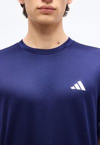 Κοντινό πλάνο ενός ατόμου που φοράει μπλε ναυτικό αθλητικό μπλουζάκι με λευκό λογότυπο Adidas στο αριστερό στήθος.