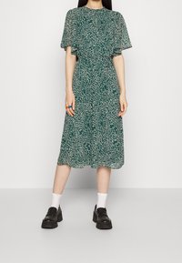 Vihreä mekko kukkakuviolla, lyhyillä liehuvilla hihoilla ja kerroksellisella muotoilulla, valmistettu kevyestä kankaasta. Yhdistetty tukeviin mustiin kenkiin.