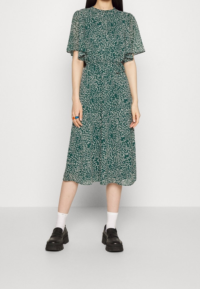 Vihreä mekko kukkakuviolla, lyhyillä liehuvilla hihoilla ja kerroksellisella muotoilulla, valmistettu kevyestä kankaasta. Yhdistetty tukeviin mustiin kenkiin.