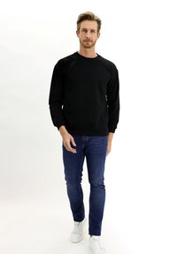Sudadera de algodón negra con un escote redondo y mangas largas raglán, combinada con jeans ajustados de color azul y zapatillas blancas.