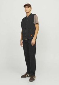 Black denim vest with zip closure, front pockets, and a brown logo patch, worn over a brown short-sleeve shirt and black pants. Brown sneakers.