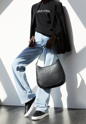 Person wearing black Calvin Klein shirt, black blazer, light blue jeans, black sneakers, holding black CK handbag against white background.