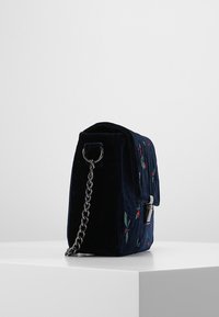 Námořnicky modrá sametová kabelka s květinovou výšivkou, obdélníkového tvaru, s řetízkovým popruhem, magnetickým zapínáním a doplňky v červené a zelené barvě.