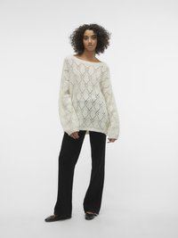 Pull en tricot crème avec des motifs en losange, manches larges et coupe loose, associé à un pantalon large noir et des chaussures plates.
