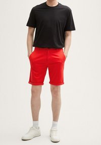 Camiseta negra de cuello redondo, pantalones cortos rojos enrollados con bolsillos laterales, usados con zapatillas blancas y calcetines blancos visibles hasta el tobillo.