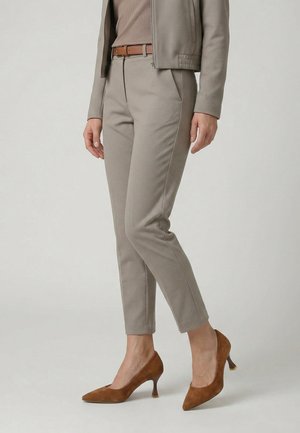 Person iført taupe slim-fit bukser, matchende jakke, lysebrunt bælte og brune ruskind høje hæle, stående mod en ensfarvet baggrund.