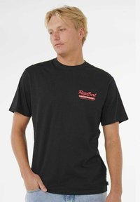 Jonge man met blond haar in een zwart Rip Curl t-shirt en lichtblauwe jeans, staand met één hand in zijn zak.