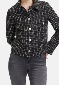 Kobieta ubrana w czarno-białą koszulę w kratę z długimi rękawami i ciemnoszare spodnie, z rękami spoczywającymi za plecami.