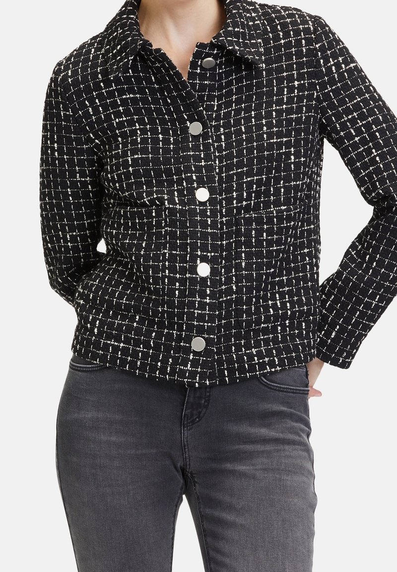 Kobieta ubrana w czarno-białą koszulę w kratę z długimi rękawami i ciemnoszare spodnie, z rękami spoczywającymi za plecami.