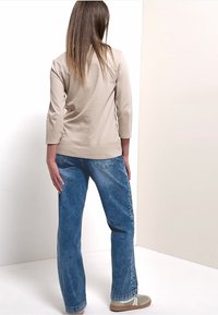 Donna in piedi con la schiena rivolta verso lo spettatore, indossa una maglietta beige a maniche lunghe, jeans blu e sneakers beige, su uno sfondo bianco e beige chiaro.