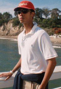 Fiatal férfi napszemüvegben, piros sapkával, fehér galléros pólóval és derekán megkötött sötétkék pulóverrel áll a korlátnál az óceán és a sziklás part közelében.