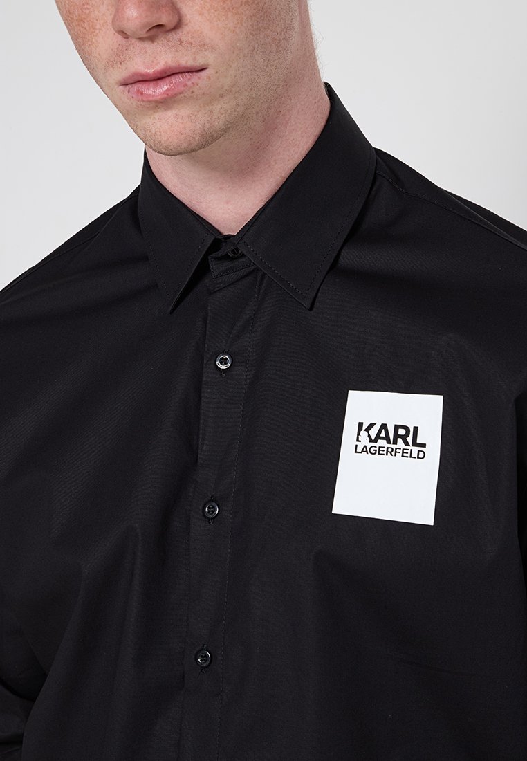 Gros plan d'une personne portant une chemise noire à boutons avec une étiquette rectangulaire blanche portant l'inscription "KARL LAGERFELD" sur la poitrine.