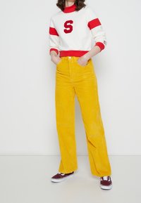 Bílá mikina se červenými pruhy a logem "S", doplněná o vysoké žluté manšestrové kalhoty. Model má na sobě vínové tenisky.