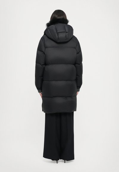Schwarzer gepolsterter langer Mantel mit Kapuze und Kunstfellbesatz, mit einem steppierten Design und gerippten Bündchen, getragen über schwarzen weiten Hosen.