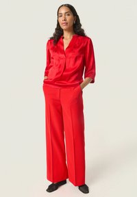 Blusa de satén roja con escote en V y mangas tres cuartos, combinada con pantalones anchos rojos. El conjunto presenta líneas elegantes y modernas.