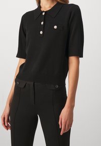 Czarny polo sweter z krótkim rękawem i kołnierzykiem, z detalami w postaci guzików z przodu oraz kieszonką na piersi. Miękka tkanina i dopasowany krój.