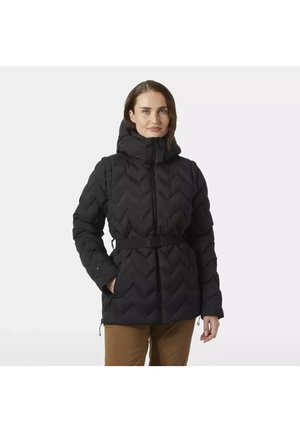 Mujer con chaqueta de invierno negra acolchada con capucha y cinturón, de pie con una mano en el bolsillo contra un fondo liso.