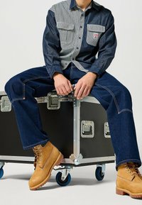 Džínová košile s pruhovanými a jednobarevnými panely, dvěma náprsními kapsami, spárovaná s tmavě modrými džíny a světlými šněrovacími botami. Sedí na černém kufříku.