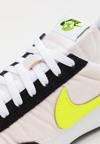 Les baskets Nike présentent une tige en tissu rose clair, des accents en daim noir, un logo jaune vif, des lacets blancs et une languette texturée et rembourrée.