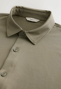 Chemise vert olive à boutons avec col pointu et étiquette de marque "NEXT" visible à l'intérieur du col.