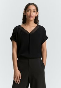 Blouse noire à manches courtes avec un col en V et un détail en mesh transparent au niveau du col. Tissu doux avec des poignets de manche retroussés. Assortie avec un pantalon noir.
