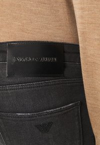 Černé džínové kalhoty z denimu s texturovanou koženou značkou s vyraženým nápisem „Emporio Armani“, vedle béžového pleteného topu.
