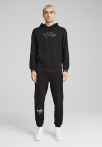 Felpa nera con logo ricamato blu, abbinata a pantaloni della tuta neri con strisce laterali turchesi e logo bianco. Realizzato in tessuto morbido.