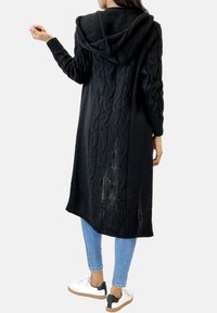 Schwarze Strickjacke mit Kapuze, mit Kabelstrickmuster, gerippten Bündchen und Saum sowie einer langen Silhouette. Getragen über blauen Jeans und Sneakers.
