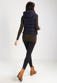 Marinblå pufferväst med huva, med horisontellt quiltade sektioner, kombinerad med en mörkgrön ribbad tröja och svarta leggings, kompletterad med bruna klackboots.