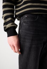 Jeans negri din denim cu un buzunar frontal simplu, purtați cu un pulover striped negru și măsliniu. Materialul pare moale, cu un ușor luciu.