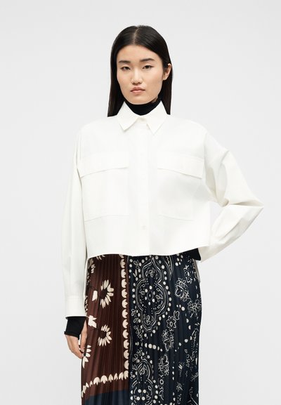 Νεαρή γυναίκα με ίσια μαύρα μαλλιά φοράει λευκό κοντό μπλουζάκι με μακριά μανίκια και πλισέ φούστα με καφέ και ναυτικό μπλε λουλουδάτα μοτίβα.