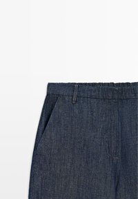 Nahaufnahme dunkelblauer Jeanshose mit elastischem Bund und Vordertaschen, dargestellt auf weißem Hintergrund.