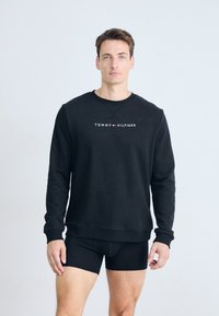 Sudadera negra con mangas largas y puños acanalados, que presenta el logo "TOMMY HILFIGER" en el centro. Llevada con pantalones cortos negros.