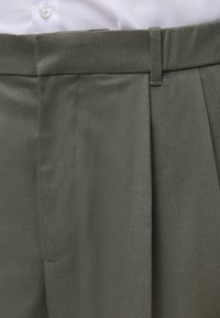 Gros plan sur la ceinture et le détail des plis d’un pantalon vert olive sur une chemise blanche avec un bouton visible.