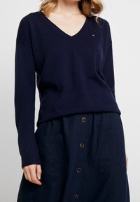 Tmavě modrý svetr s výstřihem do V z měkké látky s dlouhými rukávy. Má malý logo na hrudi a žebrované manžety, doplněný tmavou sukni na knoflíky.