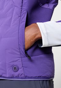 Veste isolante violette avec un design en maille texturée et une poche latérale zippée. Présente un patch logo et des détails contrastants au niveau des poignets.