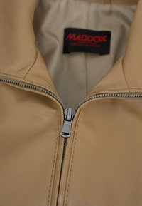 Tan leather jacket with silver zipper partially zipped, showing a black and red Maddox brand label inside the collar.