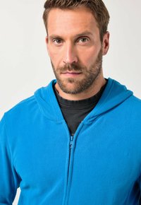 Niebieska bluza polarowa na suwak z przodu, z kapturem z troczkami. Tkanina wydaje się miękka i teksturowana. Nosi się ją na czarnej koszulce.