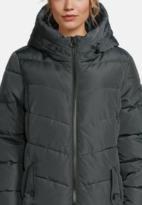 Femme portant une veste d'hiver noire matelassée avec capuche, fermeture éclair à l'avant et capuche ajustée par cordon.