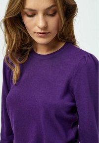 Lila gestrickter Pullover mit langen Ärmeln, rundem Halsausschnitt und leichtem Puffdetail an den Schultern. Weiche Textur und taillierter Schnitt.