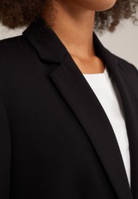 Kvinna klädd i en svart kavaj över en vit topp, som visar hals- och axelområdet med lockigt hår synligt.