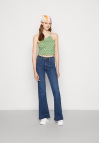 Top de flores verde com alças finas, combinado com jeans azuis aos flancos e ténis brancos. O top apresenta um design franzido.