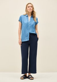 Ljust blå denimskjorta med korta ärmar, knäppning framtill; parat med marinkolvärda vida byxor; modell som bär mörka sandaler. Enfärgad bakgrund.